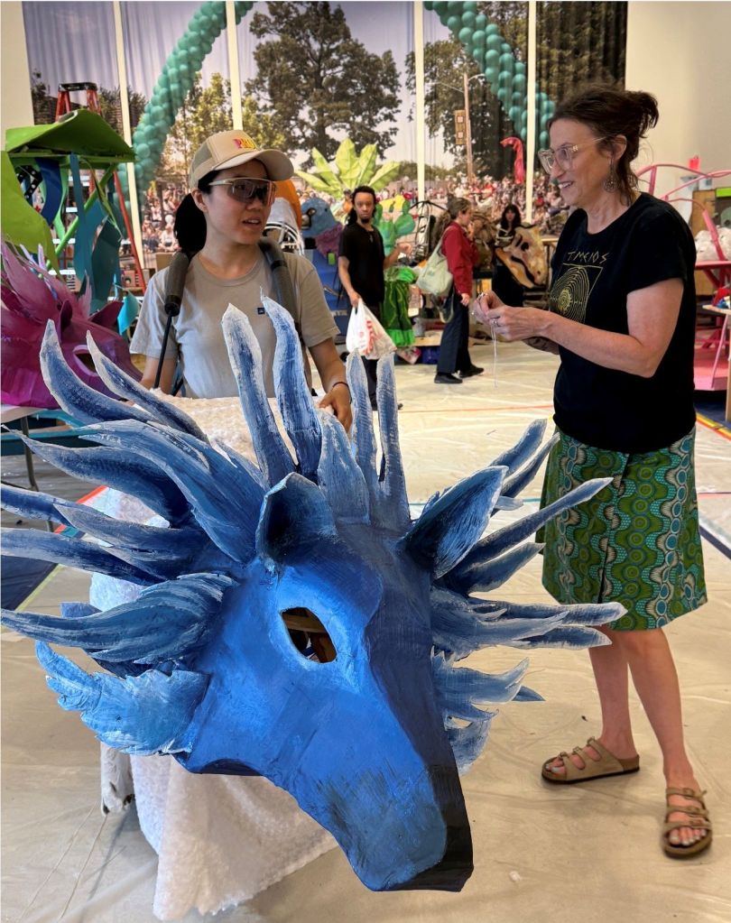 A person wearing a blue dragon costume interacts with another person at an outdoor event with colorful balloons and decorations.