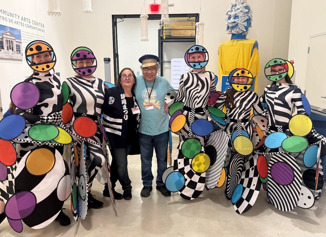 A group of people dressed in colorful, patterned costumes posing together in a community arts center. The costumes feature bold geometric shapes, stripes, and polka dots in a variety of bright colors.