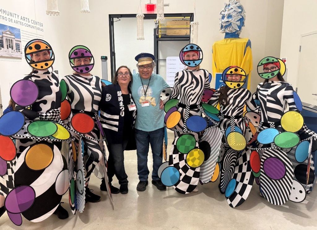 A group of people dressed in colorful, patterned costumes posing together in a community arts center. The costumes feature bold geometric shapes, stripes, and polka dots in a variety of bright colors.