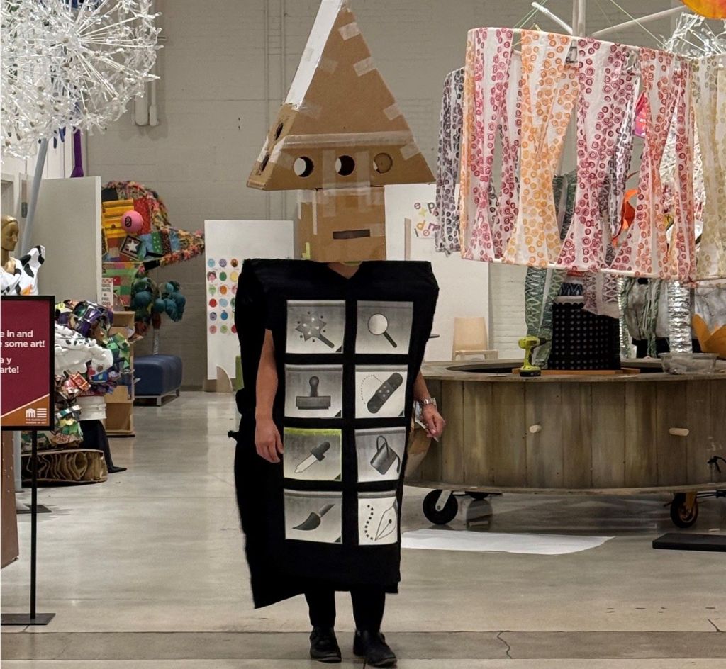 A person wearing a costume resembling a cardboard robot, standing in an art gallery or store filled with colorful artwork, sculptures, and decorations.