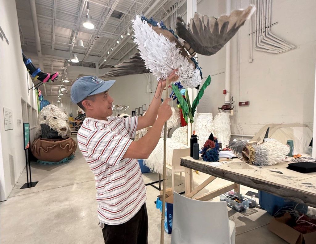 A person in a striped shirt and baseball cap is assembling a large feathered costume piece in a workshop setting with various other costume elements visible.