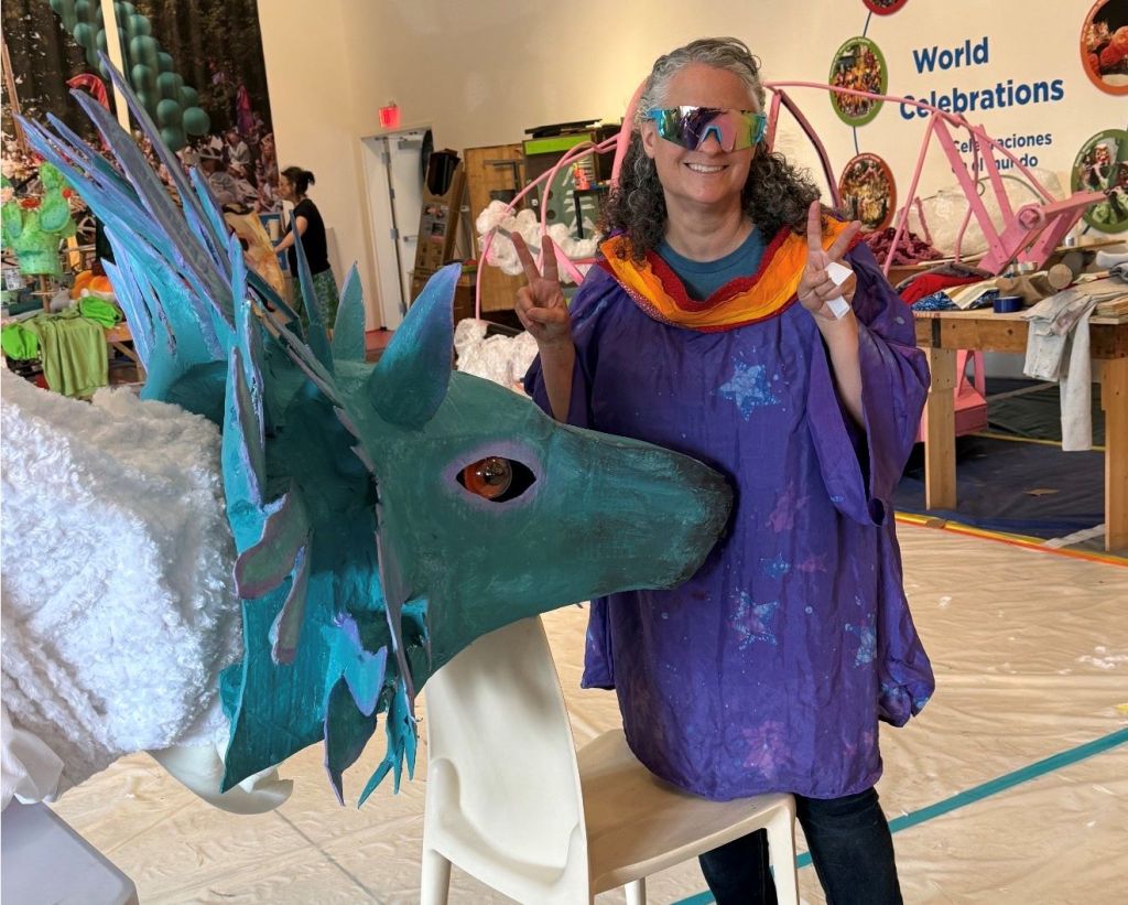 A smiling woman wearing a colorful, patterned dress and sunglasses stands next to a large, vibrant sea creature sculpture in a room decorated with world celebration-themed decor.