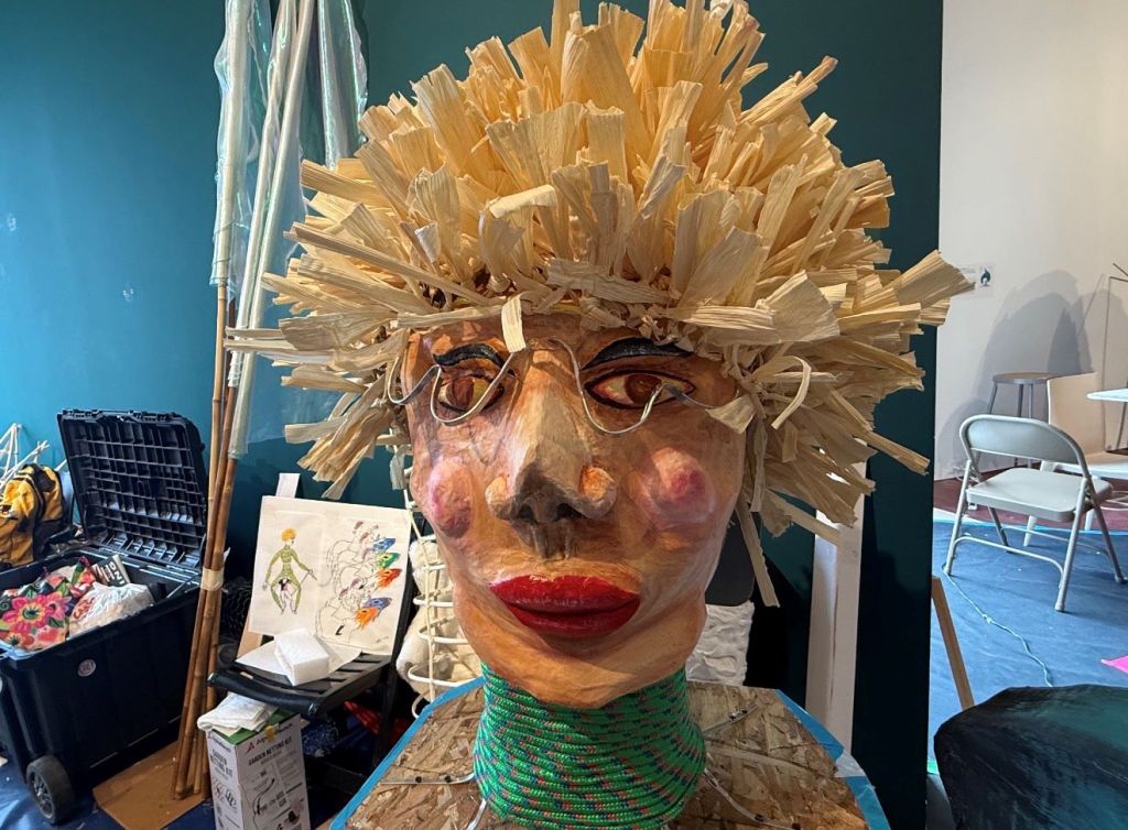 A colorful, handmade mask or headdress made of straw or dried plant material, with a human face and red lips visible through the intricate woven design.