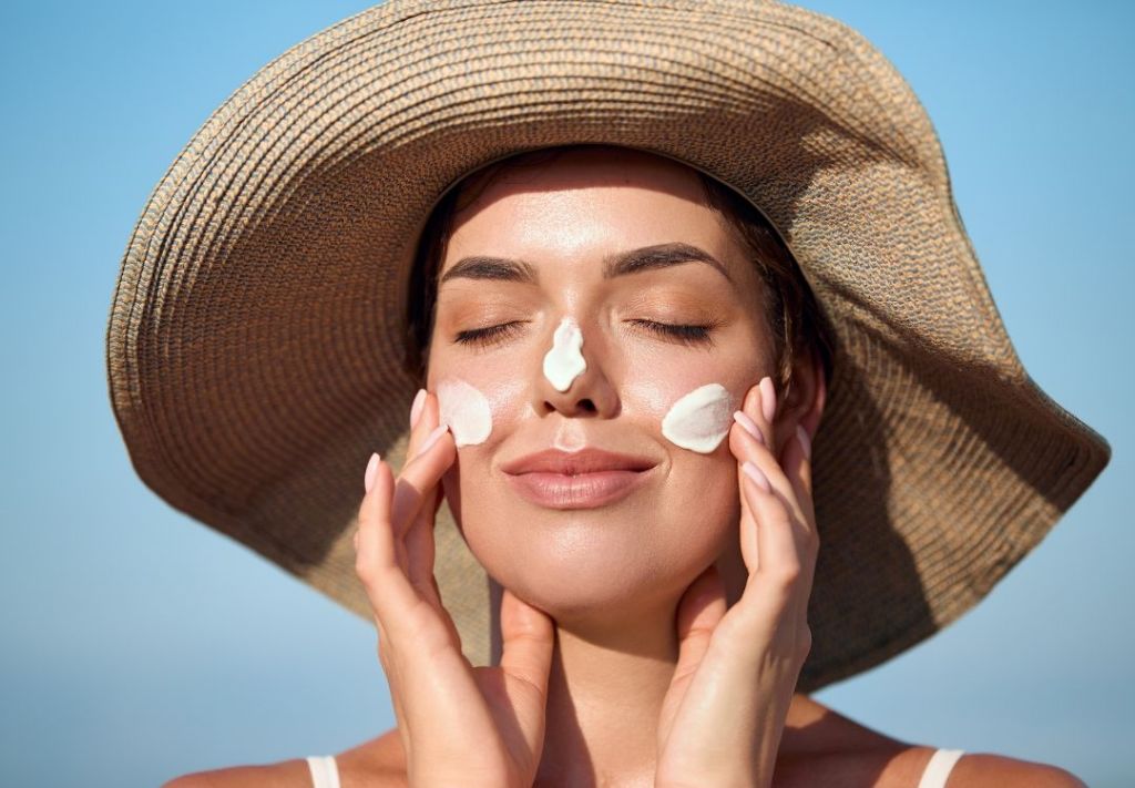 A woman in a wide-brimmed straw hat applying sunscreen to her face while enjoying the sunny outdoors.