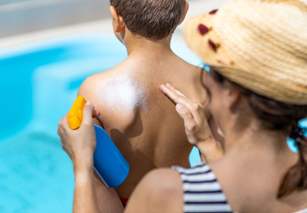 A person in a striped shirt applying sunscreen to the back of a person in a blue swimsuit by a swimming pool.