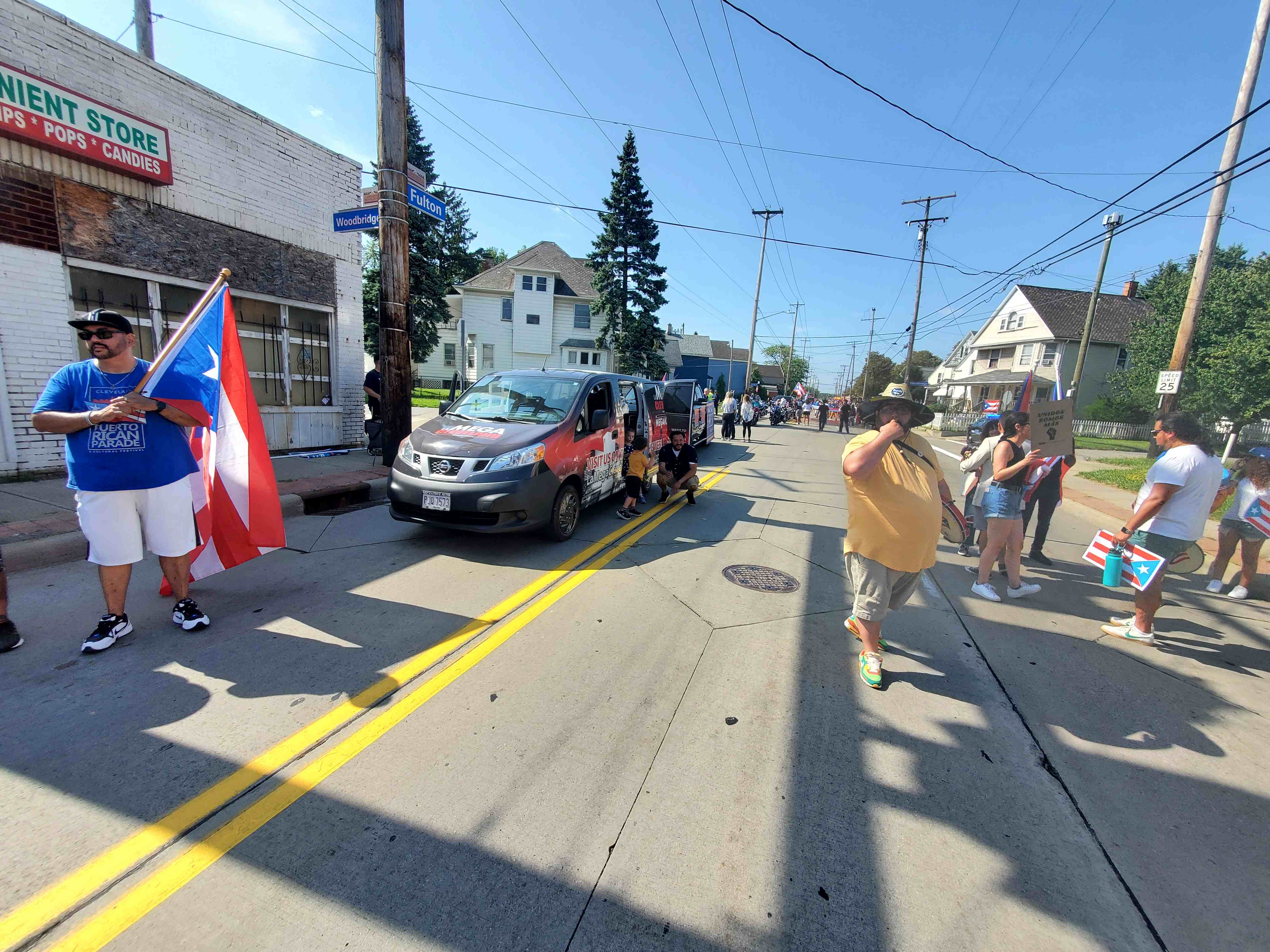 A street scene with a Puerto Rican flag, people standing on the sidewalk, and a van with "Parada Urbana" branding.
