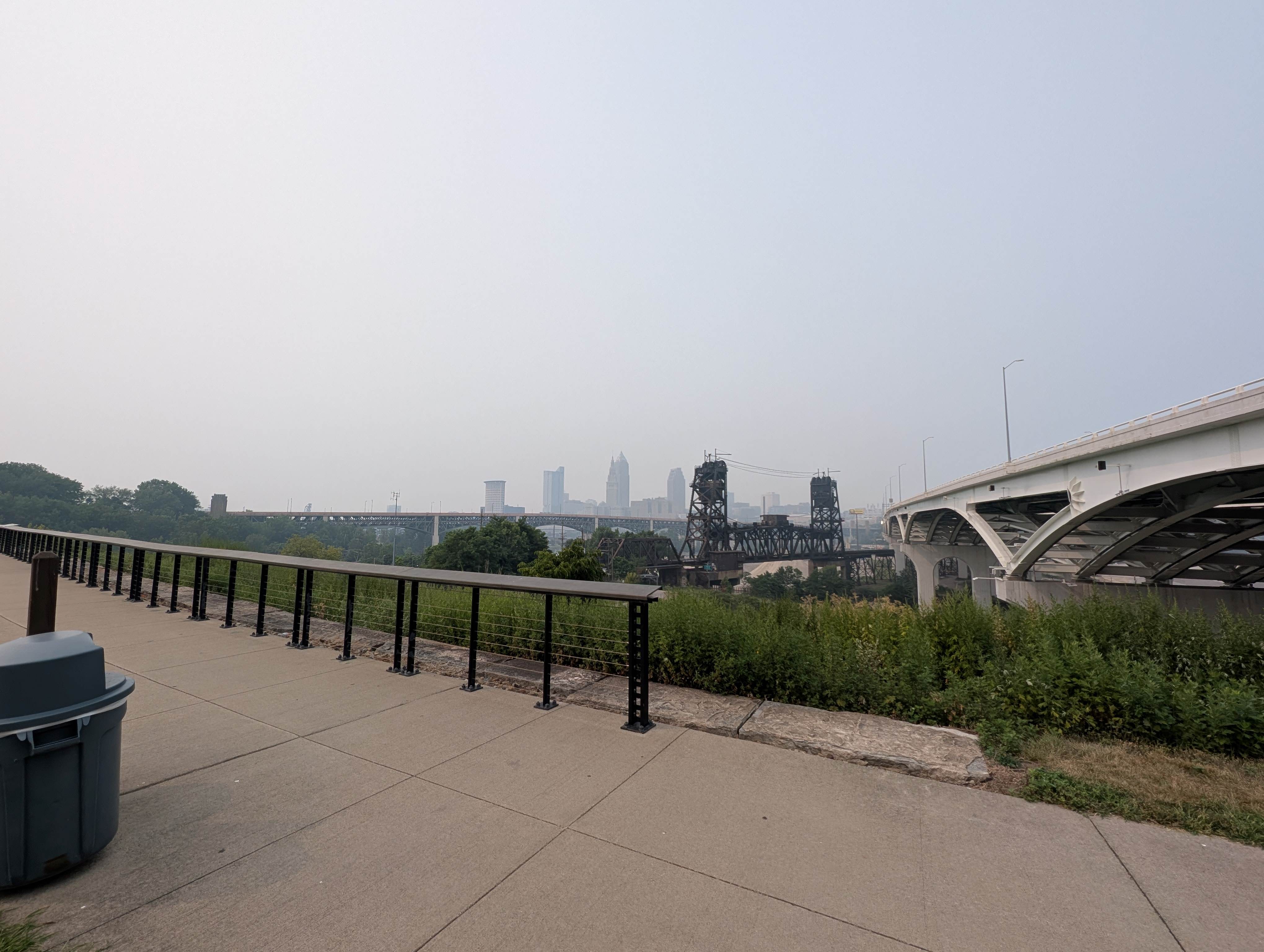 Cityscape with bridges and buildings in the distance, surrounded by grassy areas and railings.