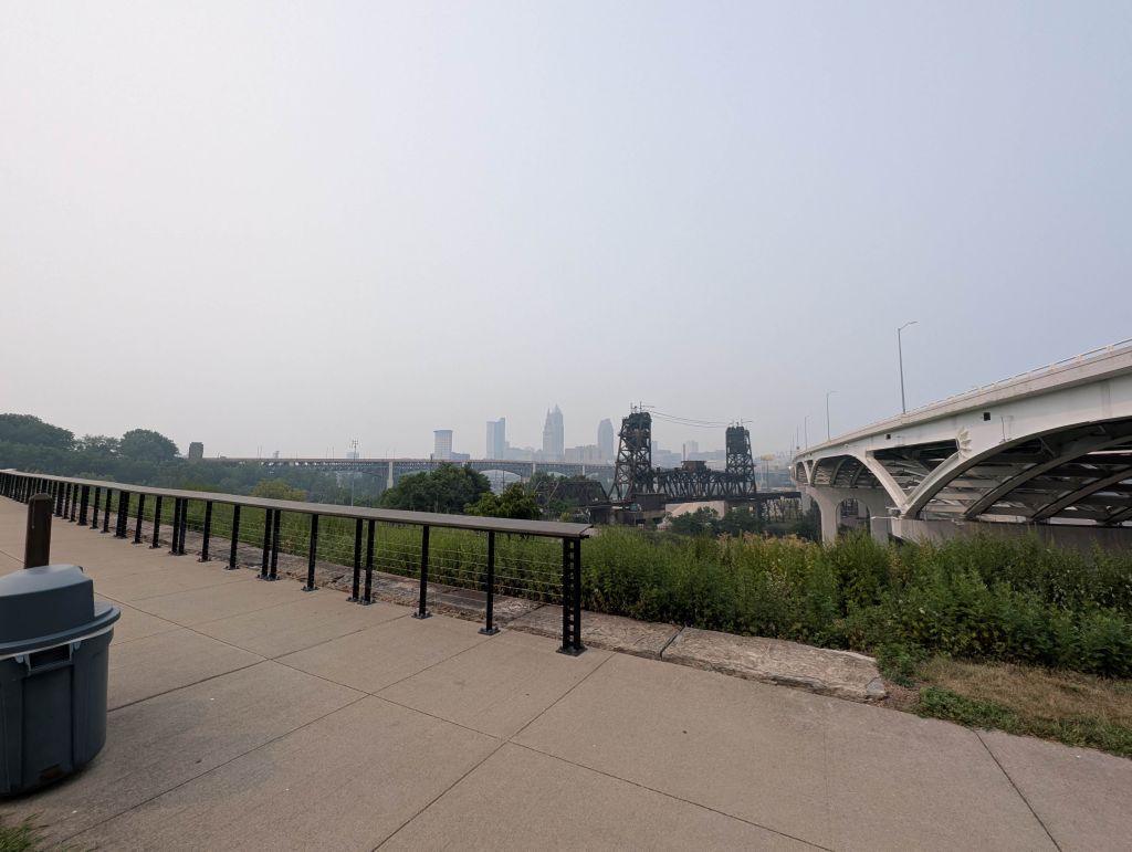 Cityscape with bridges and buildings in the distance, surrounded by grassy areas and railings.