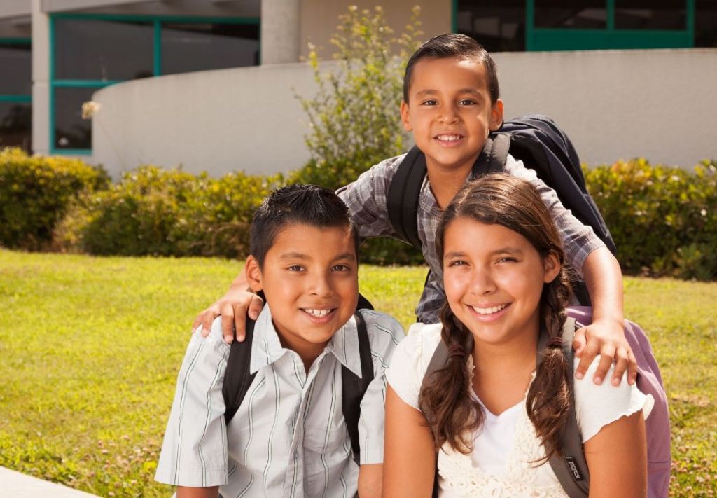 Three smiling children in school uniforms standing together outdoors in front of greenery.