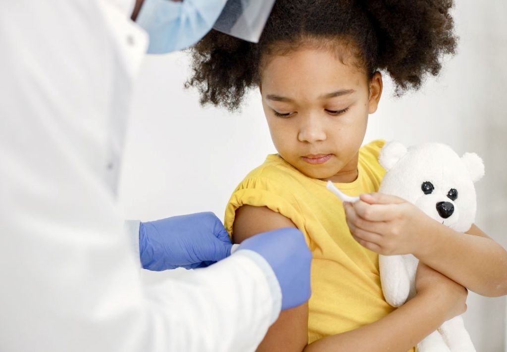A young girl in a yellow shirt holding a stuffed animal while a medical professional in blue gloves examines her.