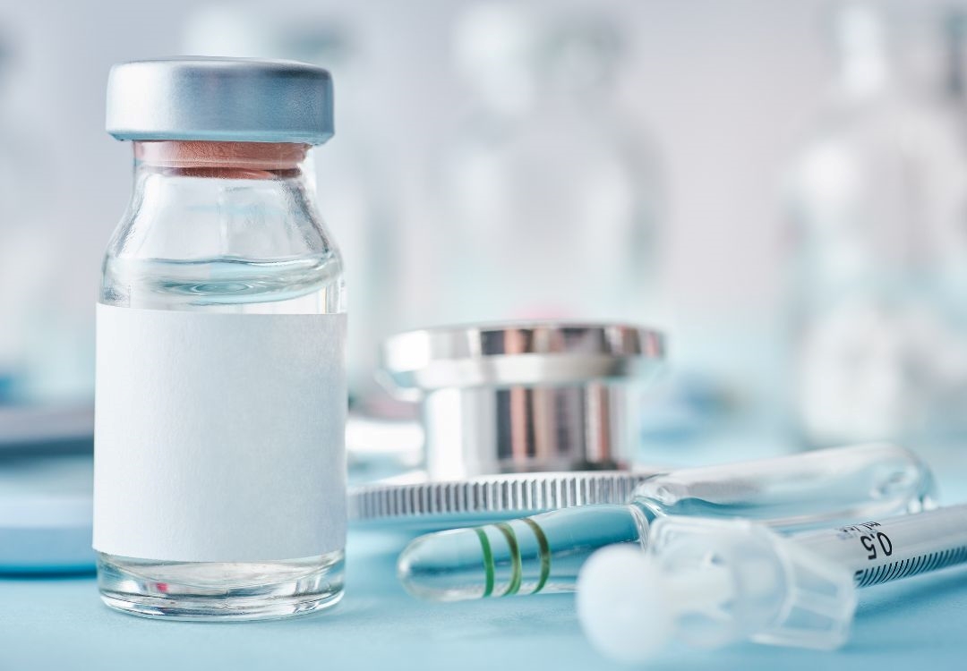 A glass vial containing a clear liquid, accompanied by a syringe and other medical equipment on a blurred background.