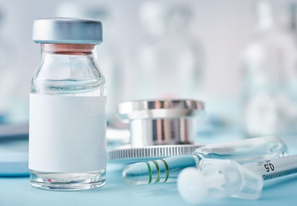 A glass vial containing a clear liquid, accompanied by a syringe and other medical equipment on a blurred background.