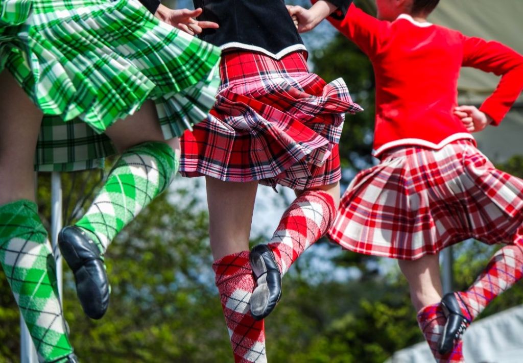 People wearing traditional Scottish tartan plaid skirts and dresses performing a traditional Scottish dance outdoors.