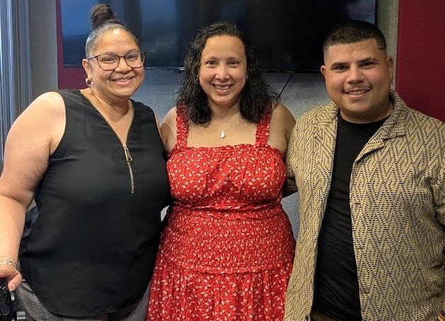 Three smiling people posing together, a woman in a red floral dress and two men in casual outfits.
