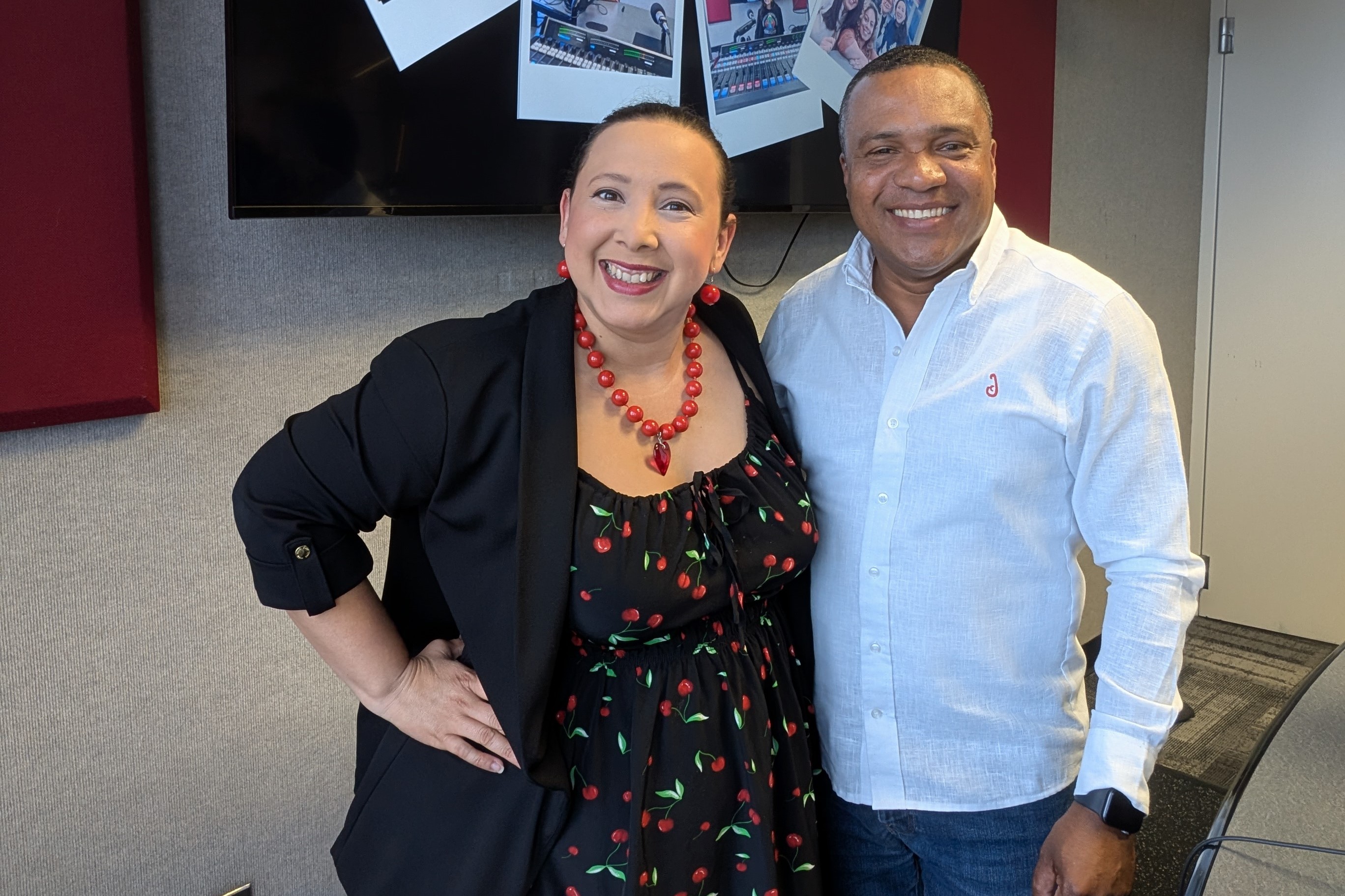Two smiling people, a woman in a black jacket and red necklace, and a man in a white shirt, posing together in an office setting with TV screens and photos on the wall behind them.