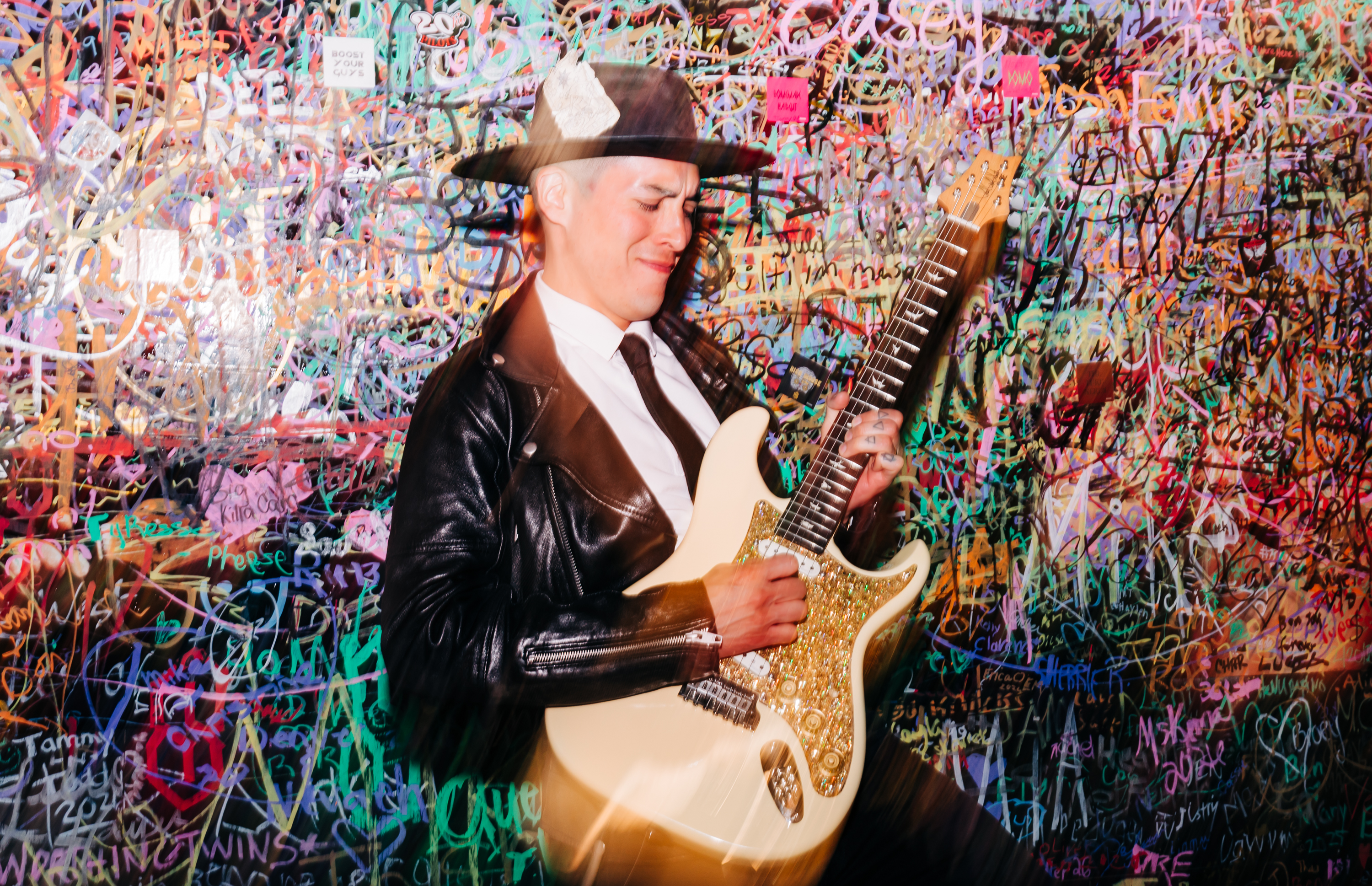 A person wearing a leather jacket and cowboy hat playing an electric guitar in front of a colorful graffiti-covered wall.