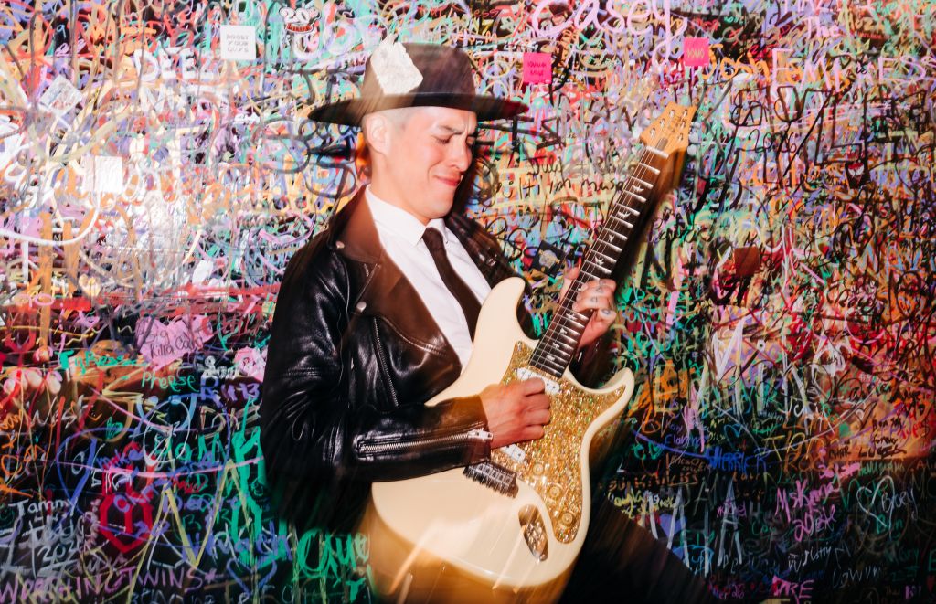 A person wearing a leather jacket and cowboy hat playing an electric guitar in front of a colorful graffiti-covered wall.