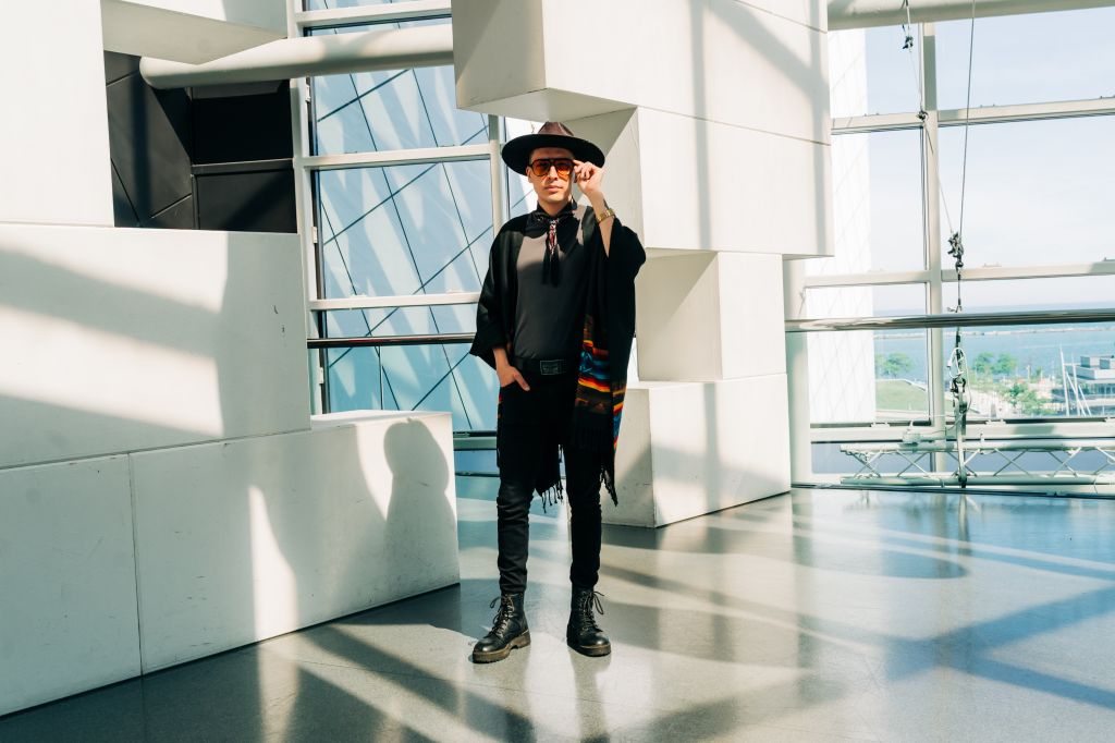 A person wearing a black hat, sunglasses, and a black outfit standing in a modern, glass-walled building.