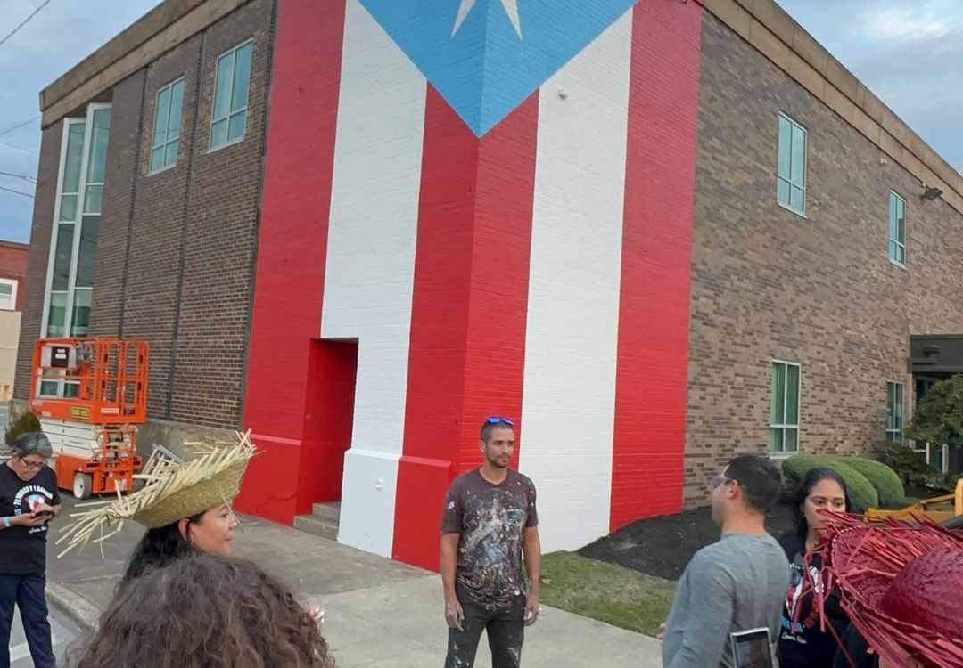 A large mural depicting the Puerto Rican flag on the side of a brick building, with several people standing in front of it.