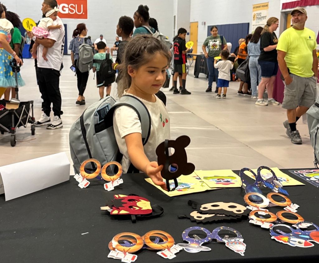 A young girl in a backpack stands at a table displaying various arts and crafts projects, including paper cutouts of eyes and other shapes, in a crowded indoor setting with people and signage in the background.