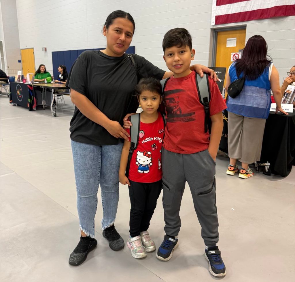 A woman and two children, a girl and a boy, standing together in a room with a red, white, and blue flag visible in the background.
