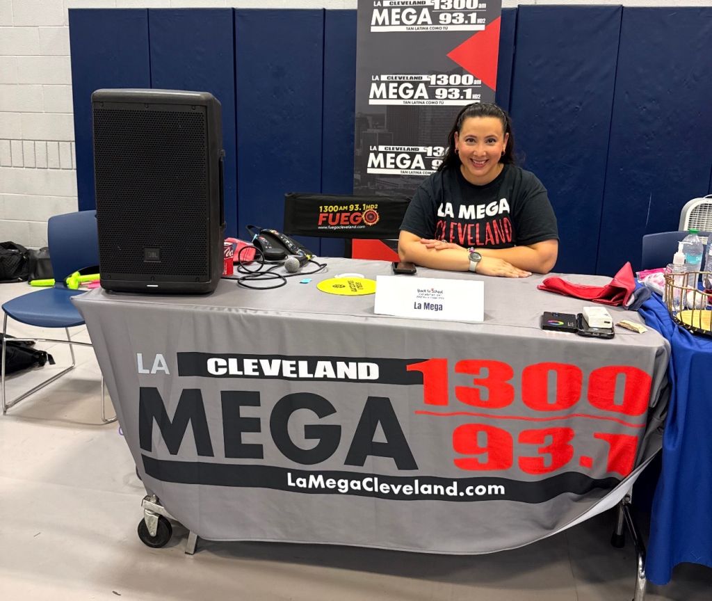 A woman smiling behind a table displaying the logo and branding for "La Cleveland Mega 1300 AM 93.7 FM". The table has various radio equipment and promotional items.