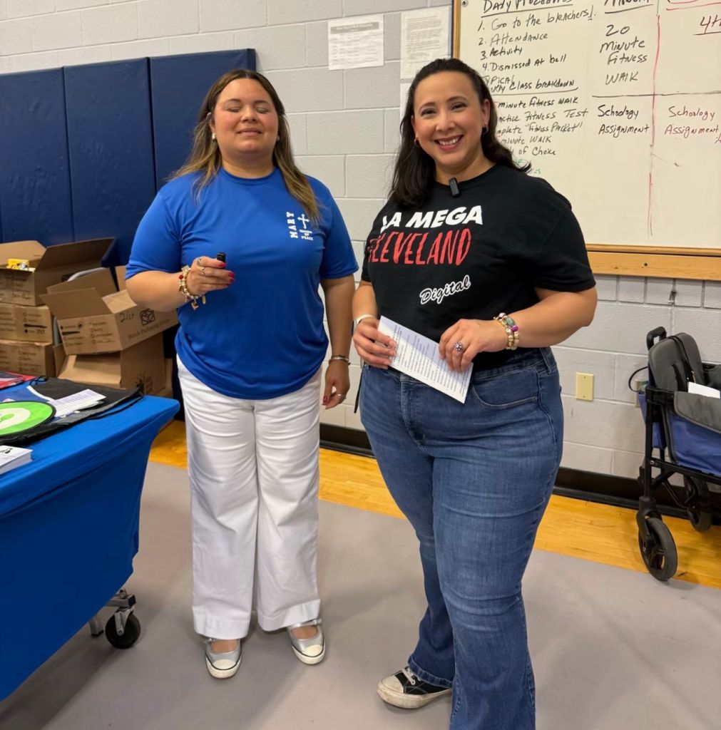Two women standing in a classroom, one wearing a blue "Mega Hit" t-shirt and the other wearing a black "La Mega Cleveland" t-shirt, both smiling.