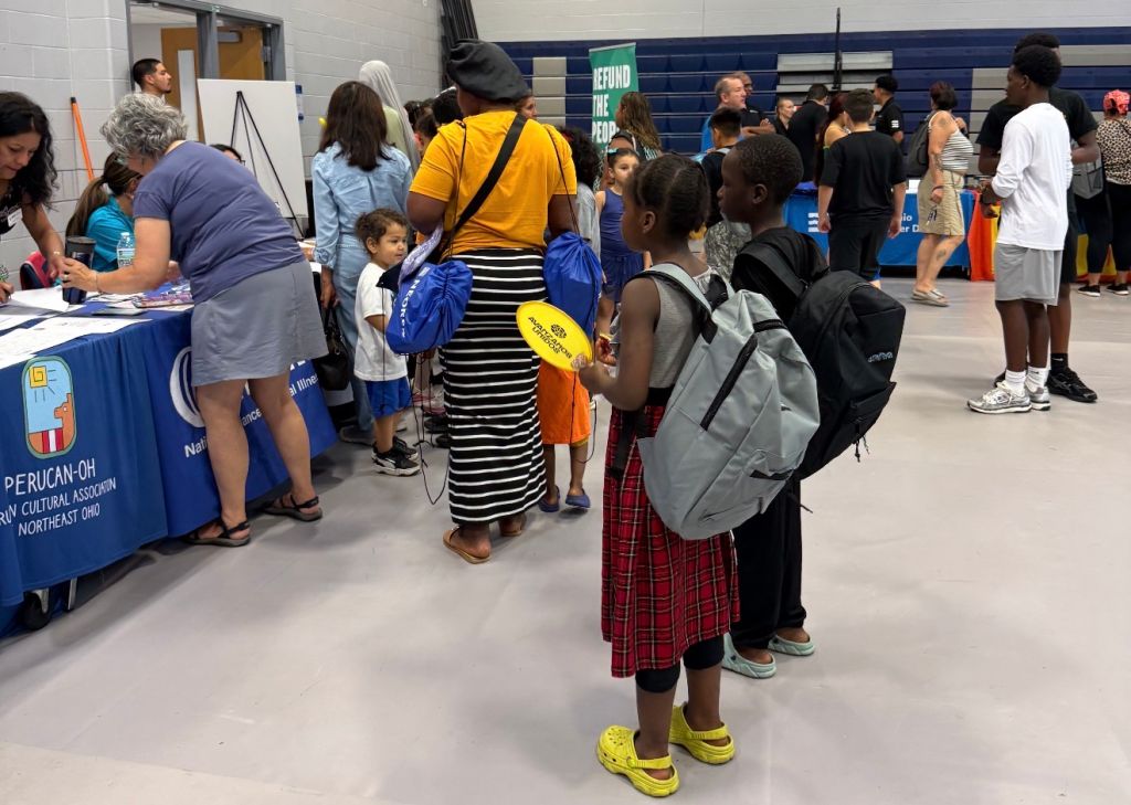 A crowded indoor event with people of various ages and backgrounds, some wearing backpacks, gathered around tables with signs and displays. The text "REFUND THE FEES" is visible on a sign.