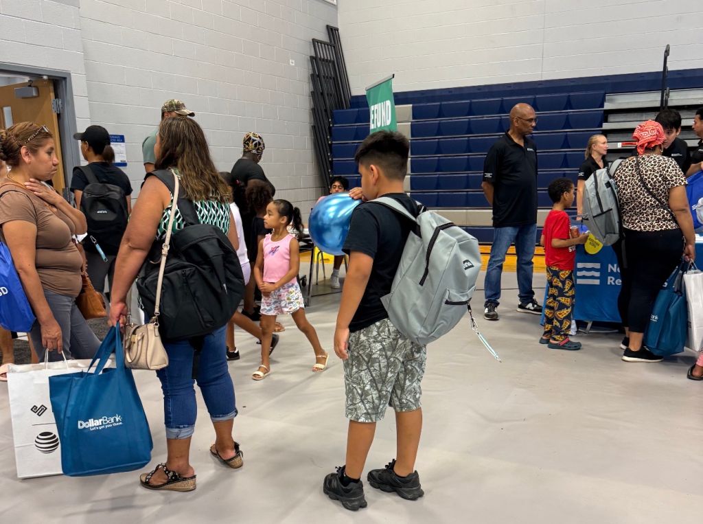 A group of people, including children, standing in a school hallway with backpacks and bags. The walls display "Gym" signage.
