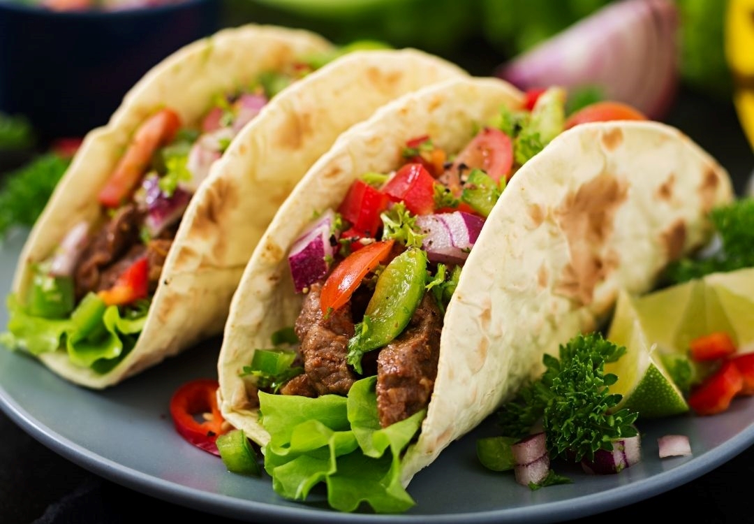 Plate of three tacos filled with meat, lettuce, tomatoes, and other vegetables, served on a green plate with lime wedges.