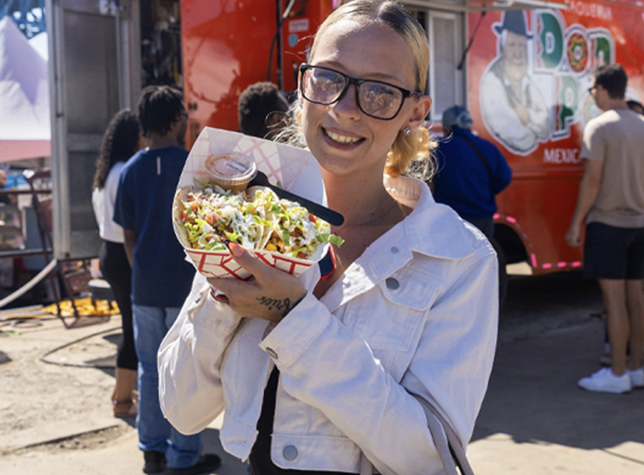 A smiling woman in a white jacket holding a burrito or wrap in front of a colorful storefront.